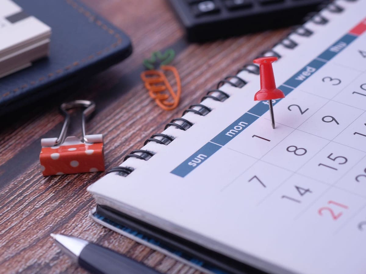 Eine rote Reißzwecke steckt auf einem Tischkalender neben Notizbuch und Büroklammern.