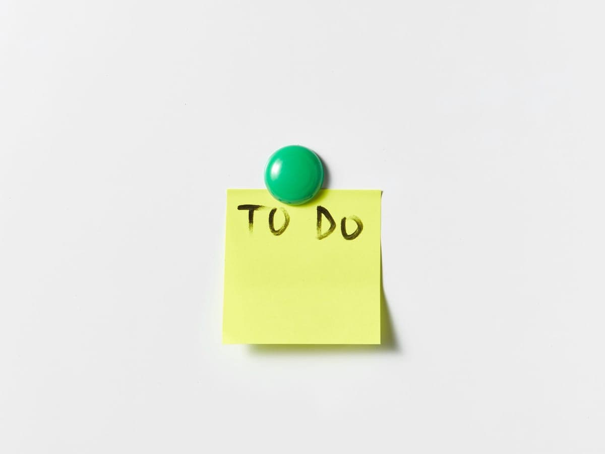 Ein gelber Notizzettel mit der Aufschrift „TO DO", mit einem Magneten an eine helle Fläche geheftet.